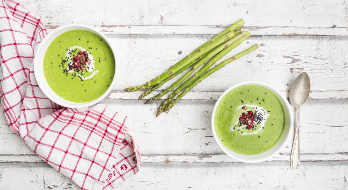 Untergrund aus Holz stehen zwei Schalen. In jeder Schale ist Spargel-Suppe mit Granatapfelkernen und Sesam-Topping / Handtuch und  Löffel / Draufsicht