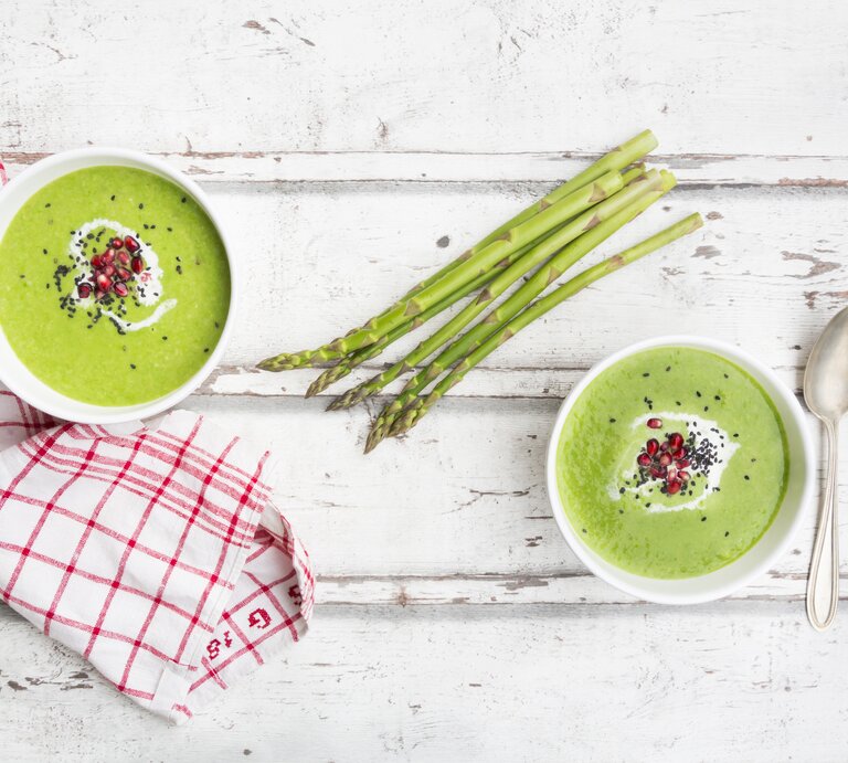 Untergrund aus Holz stehen zwei Schalen. In jeder Schale ist Spargel-Suppe mit Granatapfelkernen und Sesam-Topping / Handtuch und  Löffel / Draufsicht | © Getty Images / Westend61