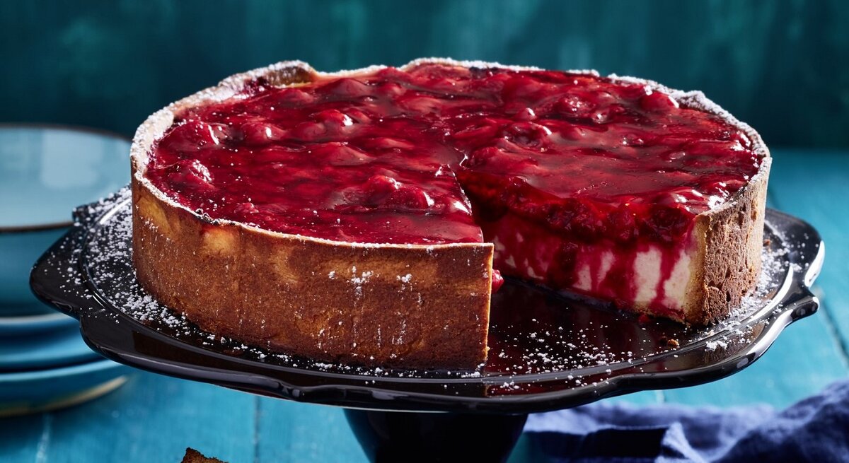 Auf einem Tortenständer steht eine Schmandtorte mit Beeren-Topping. Sie ist angeschnitten und man erkennt die cremige Füllung. 