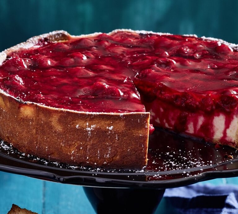 Auf einem Tortenständer steht eine Schmandtorte mit Beeren-Topping. Sie ist angeschnitten und man erkennt die cremige Füllung.  | © EAT CLUB / Isabell Triemer