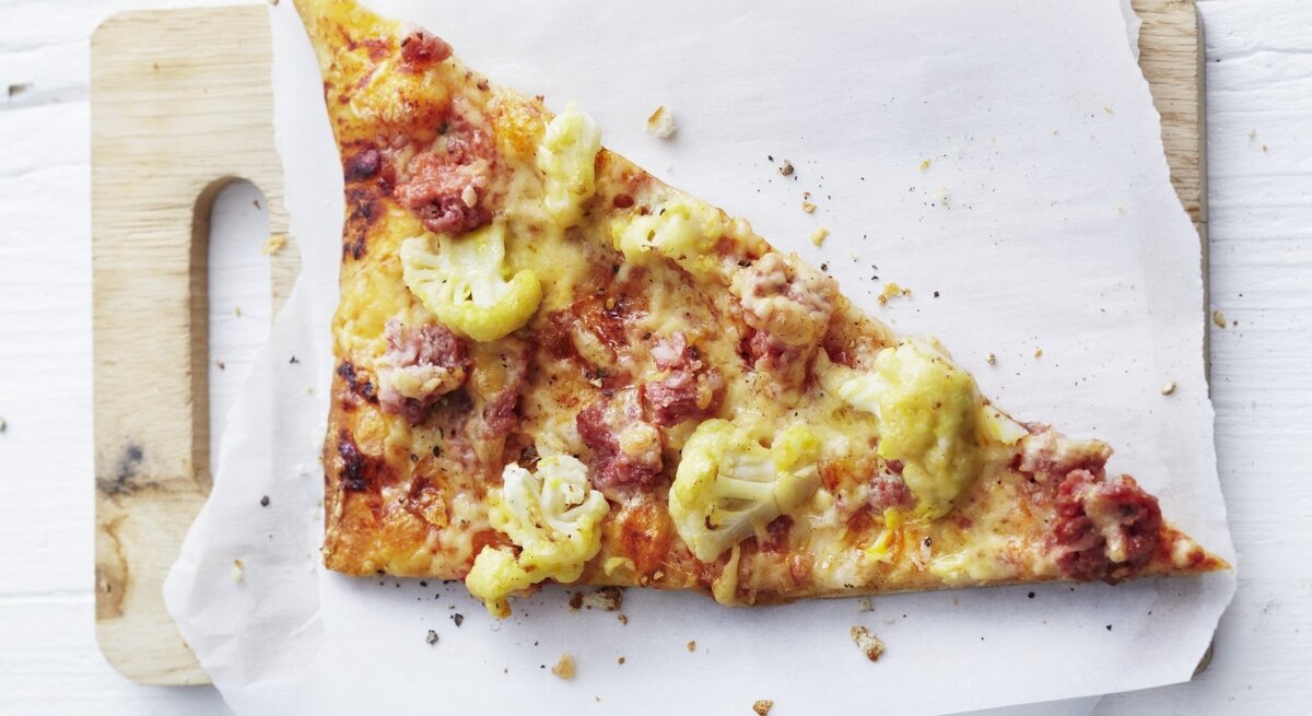 Auf einem Schneidebrett liegt ein Stück Pizza auf norddeutsche Art mit Blumenkohl und Mettwurst. Sie ist frisch gebacken, goldbraun und direkt von oben fotografiert. 