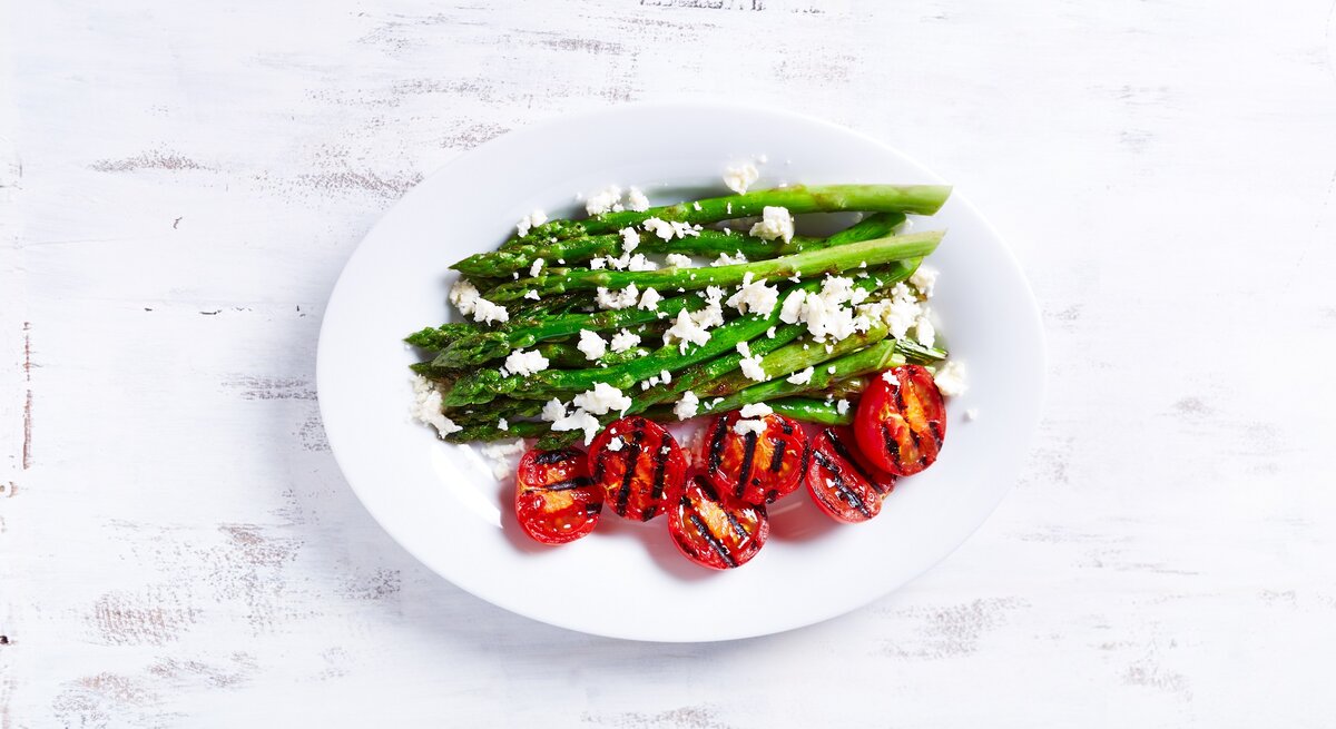 Ein Teller gegrillter Spargel mit Zitrone und Feta sowie gegrillten Tomaten in der Draufsicht.