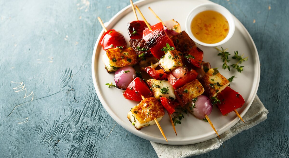 4 Tofu-Grillspieße mit Gemüse auf einem Teller, auf dem auch eine Schale mit Dip steht. 