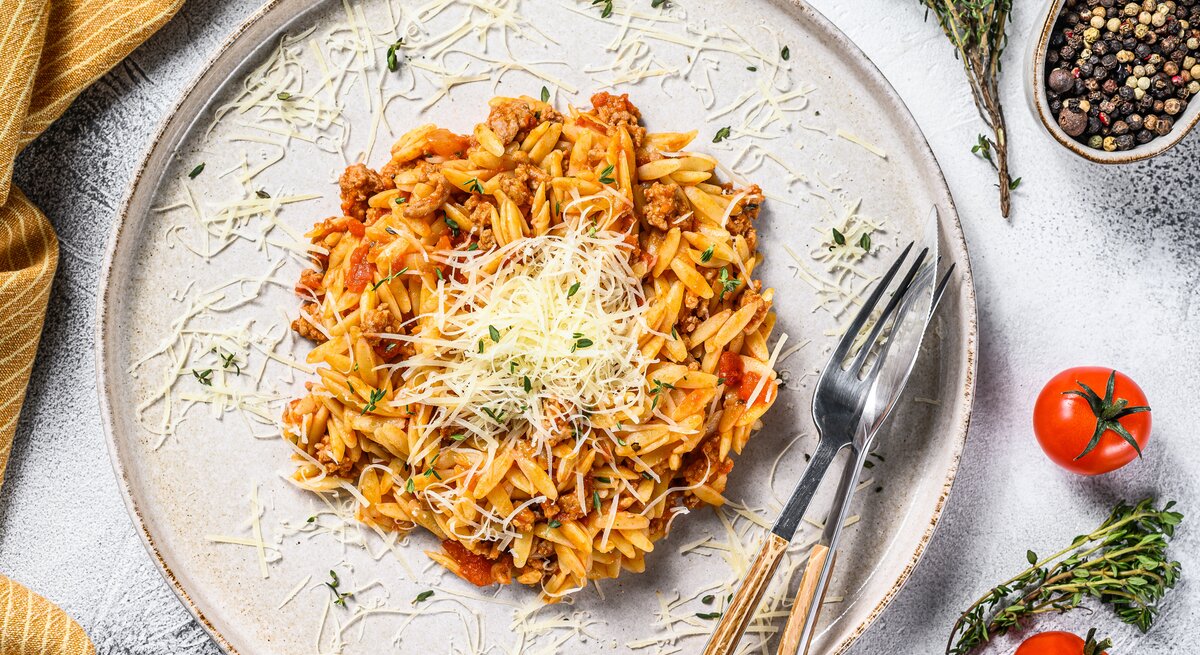 Ein Teller veganes Orzorisotto in der Draufsicht, mit veganem Parmesan bestreut, daneben Besteckt, ein Geschirrtuch, Tomaten und Gewürze.