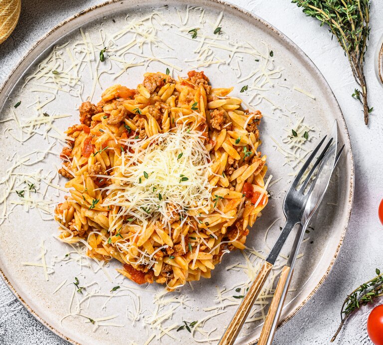 Ein Teller veganes Orzorisotto in der Draufsicht, mit veganem Parmesan bestreut, daneben Besteckt, ein Geschirrtuch, Tomaten und Gewürze. | © Getty Images/ Vladimir Mironov