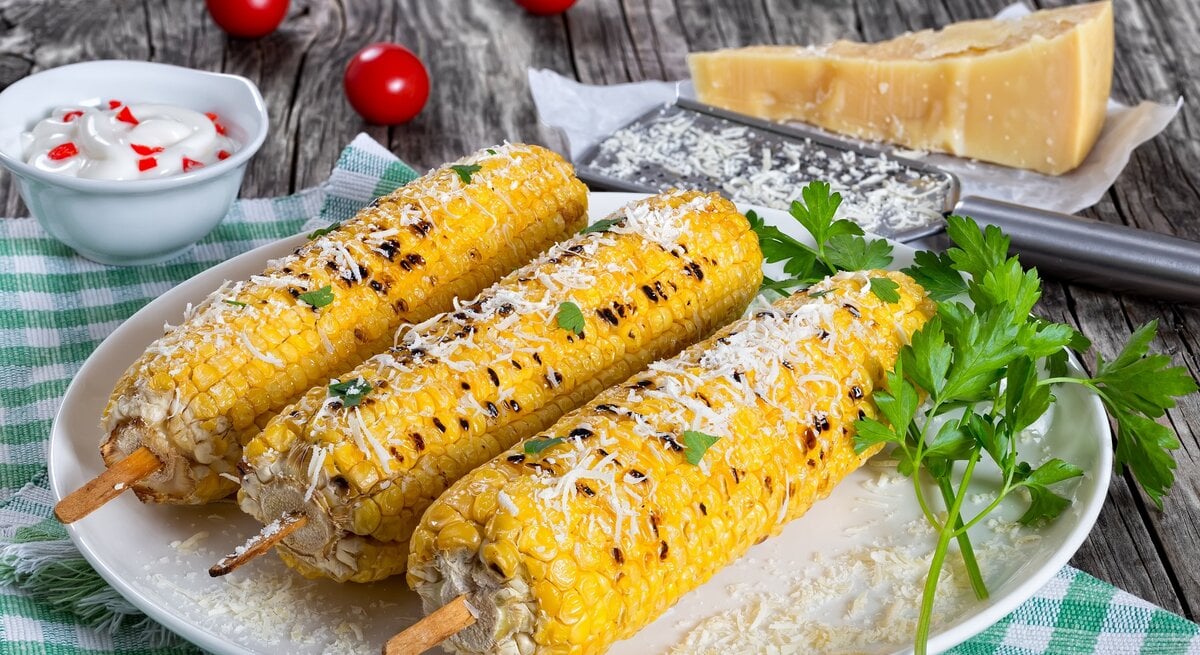 Drei Maiskolben mit Parmesan auf einem weißen Teller seitlich auf einem Holztisch fotografiert. Alles auf einer grün-weiß-karierten Tischdecke mit Parmesan und Reibe im Hintergrund. 