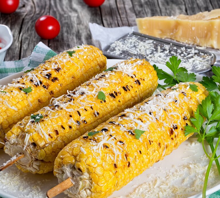 Drei Maiskolben mit Parmesan auf einem weißen Teller seitlich auf einem Holztisch fotografiert. Alles auf einer grün-weiß-karierten Tischdecke mit Parmesan und Reibe im Hintergrund.  | © Shutterstock/ from my point of view