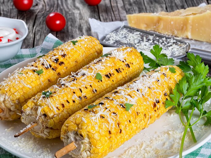 Drei Maiskolben mit Parmesan auf einem weißen Teller seitlich auf einem Holztisch fotografiert. Alles auf einer grün-weiß-karierten Tischdecke mit Parmesan und Reibe im Hintergrund.  | © Shutterstock/ from my point of view