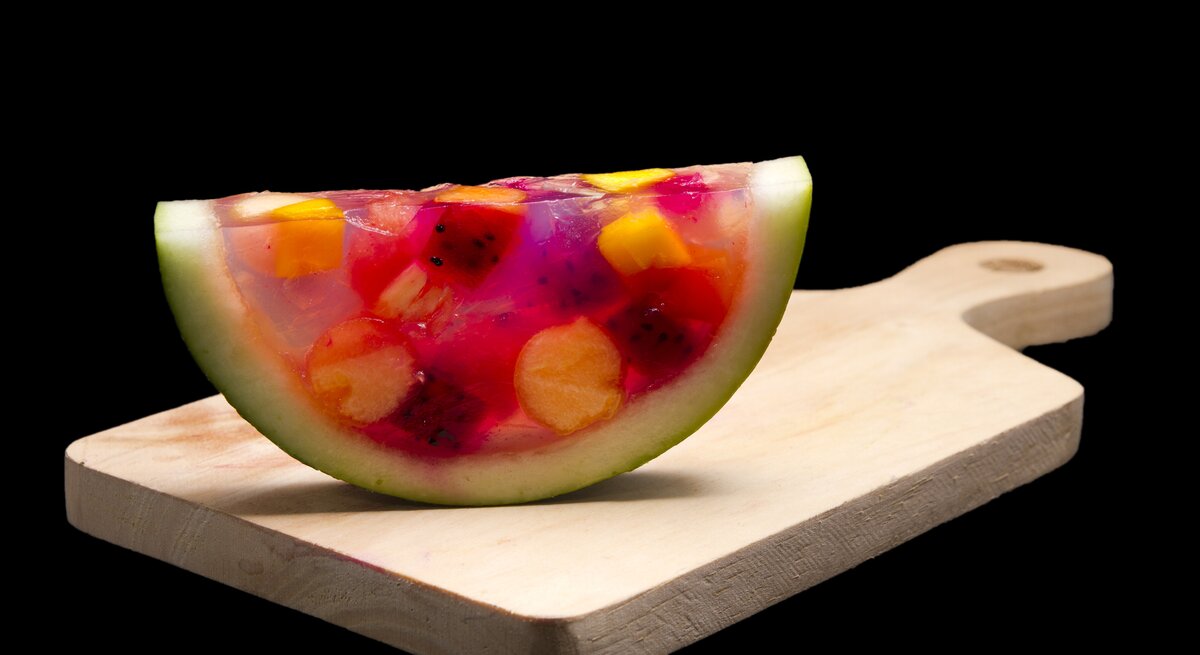 Ein Stück Watermelon-Jelly steht aufrecht auf einem braunen Holzbrett mit Griff. Das Jelly ist gefüllt mit verschiedenen Obststücken. Der Untergrund und der Hintergrund sind schwarz.