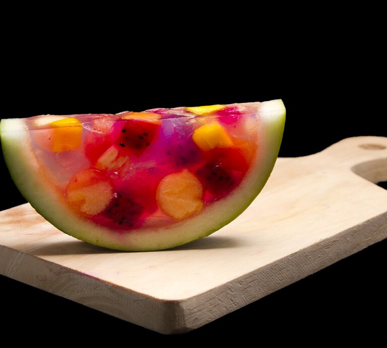 Ein Stück Watermelon-Jelly steht aufrecht auf einem braunen Holzbrett mit Griff. Das Jelly ist gefüllt mit verschiedenen Obststücken. Der Untergrund und der Hintergrund sind schwarz. | © GettyImages / halimqomarudin