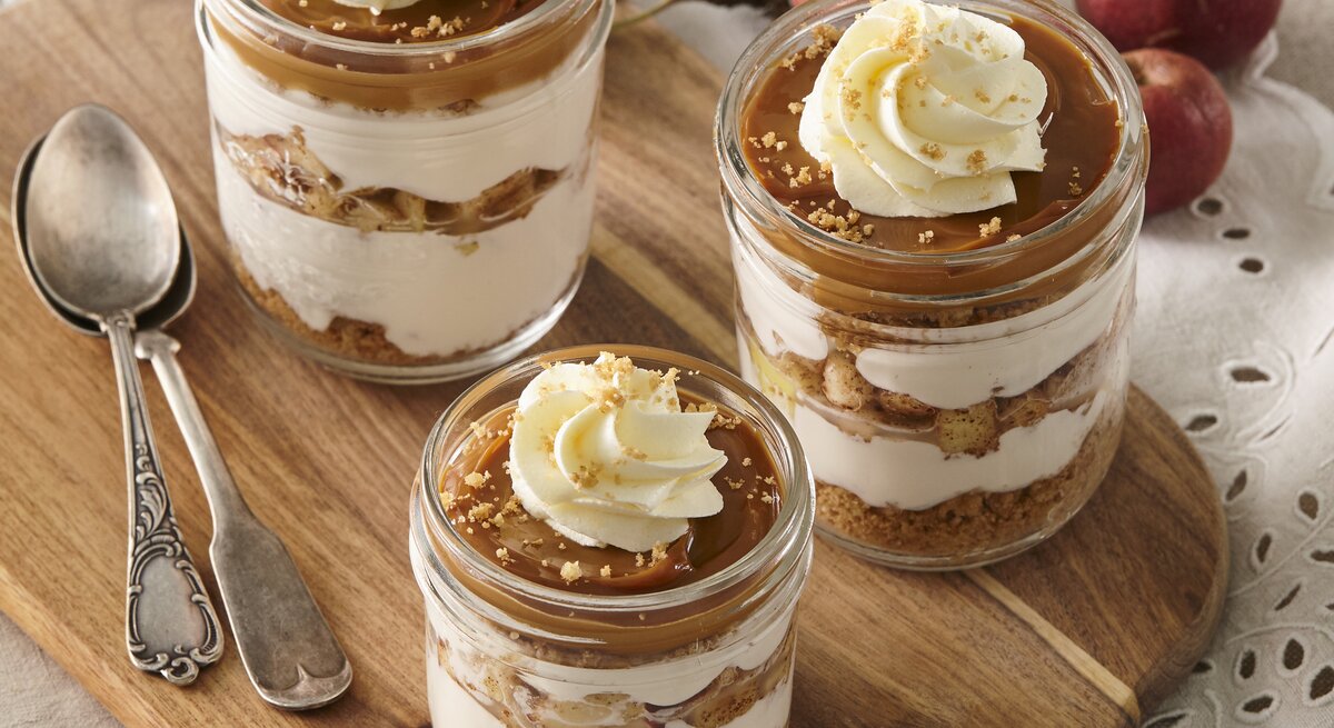 Auf einem Holzbrett stehen vier Gläser Käsekuchen im Glas mit Karamell und Apfel. Sie sind verziert mit Sahne und Keksen. Daneben liegen zwei Löffel. 