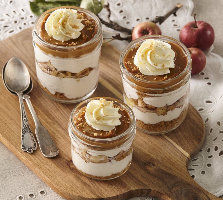 Auf einem Holzbrett stehen vier Gläser Käsekuchen im Glas mit Karamell und Apfel. Sie sind verziert mit Sahne und Keksen. Daneben liegen zwei Löffel.  | © Stockfood/Profimedia
