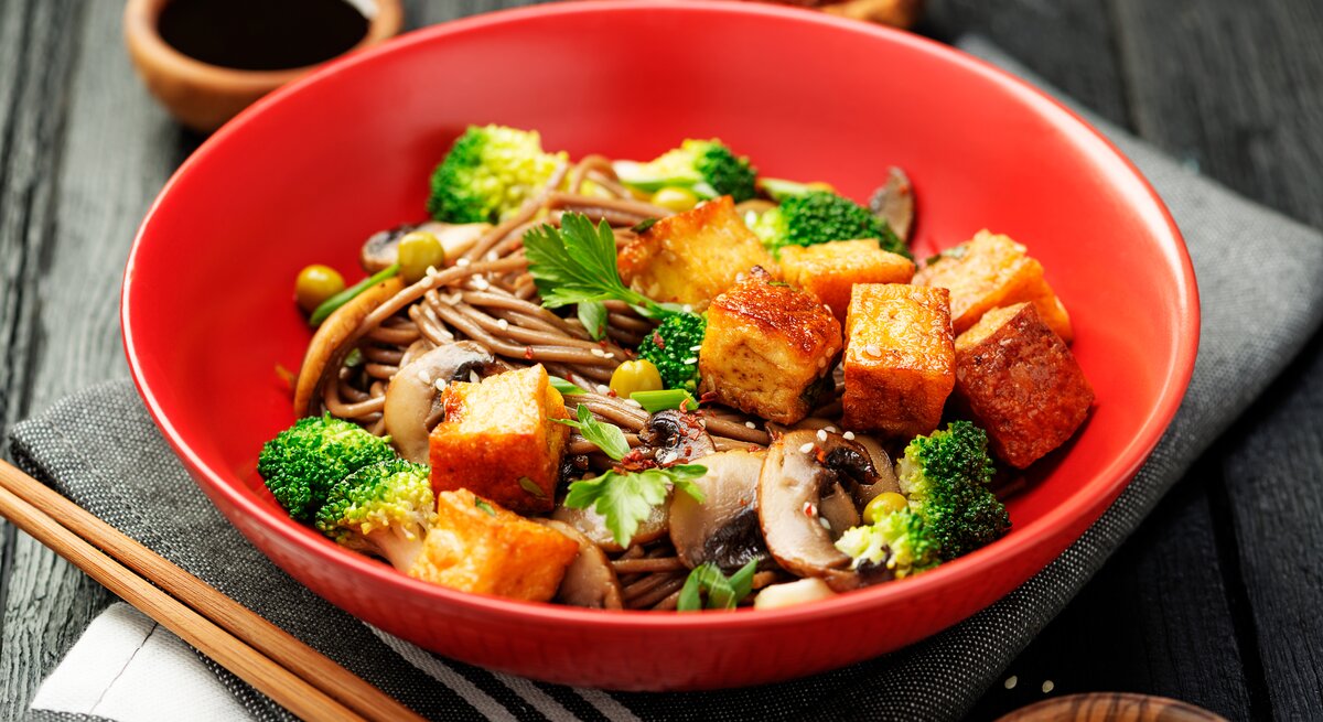 Ein tiefer roter Teller mit Soba-Nudelsalat mit Tofu und Brokkoli auf einem grauen Küchentuch.
