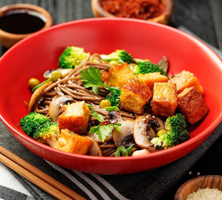 Ein tiefer roter Teller mit Soba-Nudelsalat mit Tofu und Brokkoli auf einem grauen Küchentuch. | © Shutterstock/nelea33