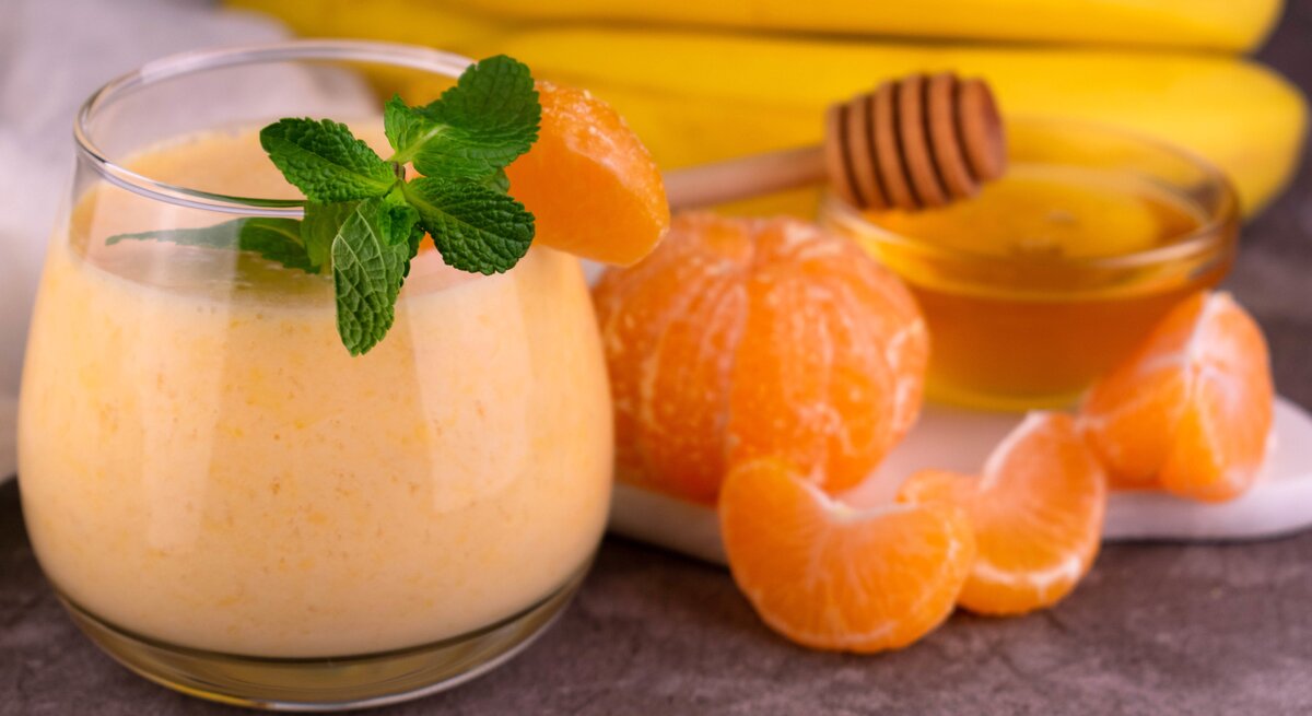 Ein Glas mit dem Smoothie. Daneben Mandarinen und Bananen sowie eine kleine Glasschale mit Honig.