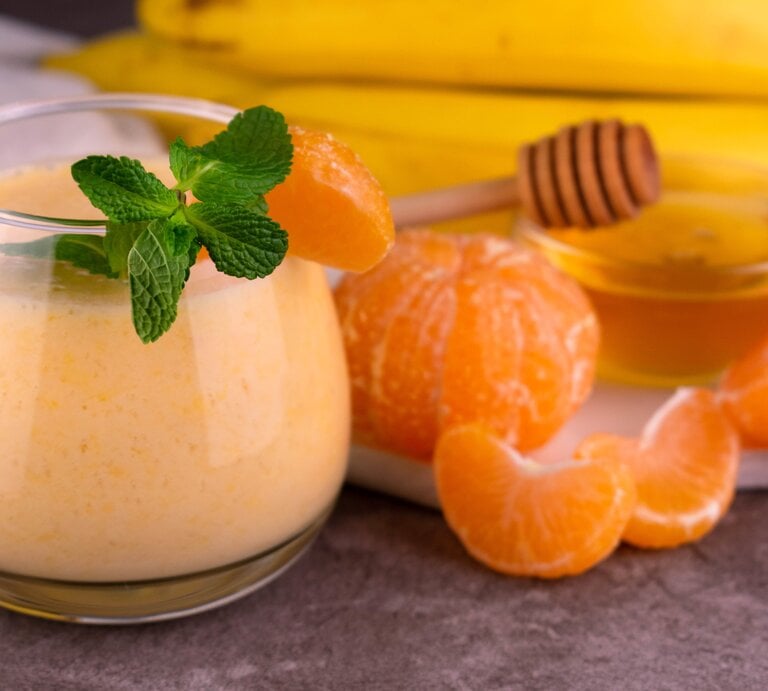 Ein Glas mit dem Smoothie. Daneben Mandarinen und Bananen sowie eine kleine Glasschale mit Honig. | © Shutterstock/ Olivka888