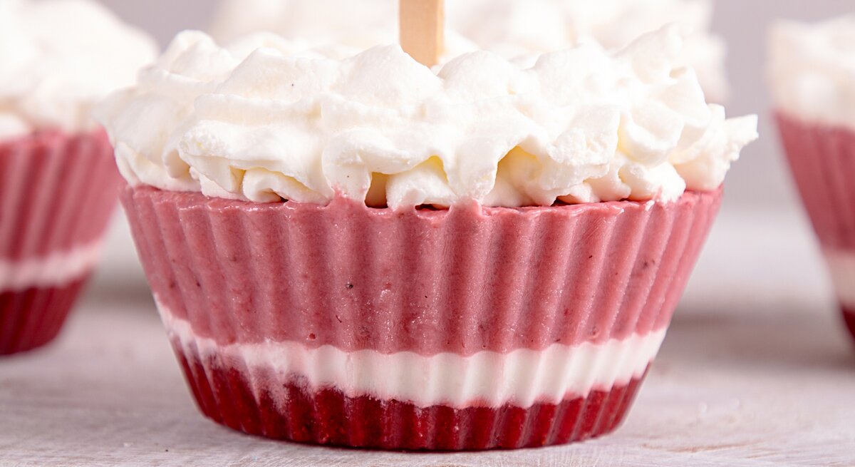Ein Frozen-Yogurt-Muffin mit drei verschiedenen Schichten, mit Schlagsahne dekoriert, im Hintergrund unscharf und angeschnitten weitere Muffins.