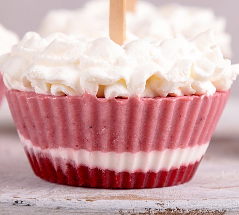 Ein Frozen-Yogurt-Muffin mit drei verschiedenen Schichten, mit Schlagsahne dekoriert, im Hintergrund unscharf und angeschnitten weitere Muffins. | © Adobe Stock/ twomeerkats