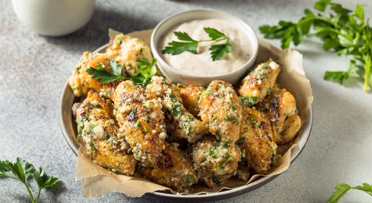 Eine Schale mit gegrillten Parmesan Chicken Wings auf hellem Untergrund mit Petersiliengarnitur und einer Schale hellem Dip. Daneben ein Bund Petersilie. Oben Pfeffermühle. Aufnahme aus der Vogelperspektive.