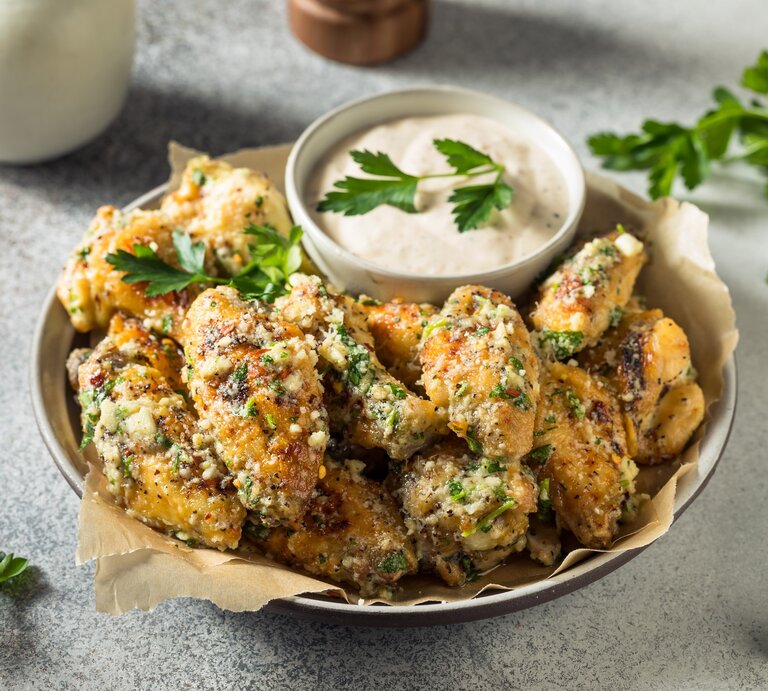 Eine Schale mit gegrillten Parmesan Chicken Wings auf hellem Untergrund mit Petersiliengarnitur und einer Schale hellem Dip. Daneben ein Bund Petersilie. Oben Pfeffermühle. Aufnahme aus der Vogelperspektive. | © Getty Images / bhofack2