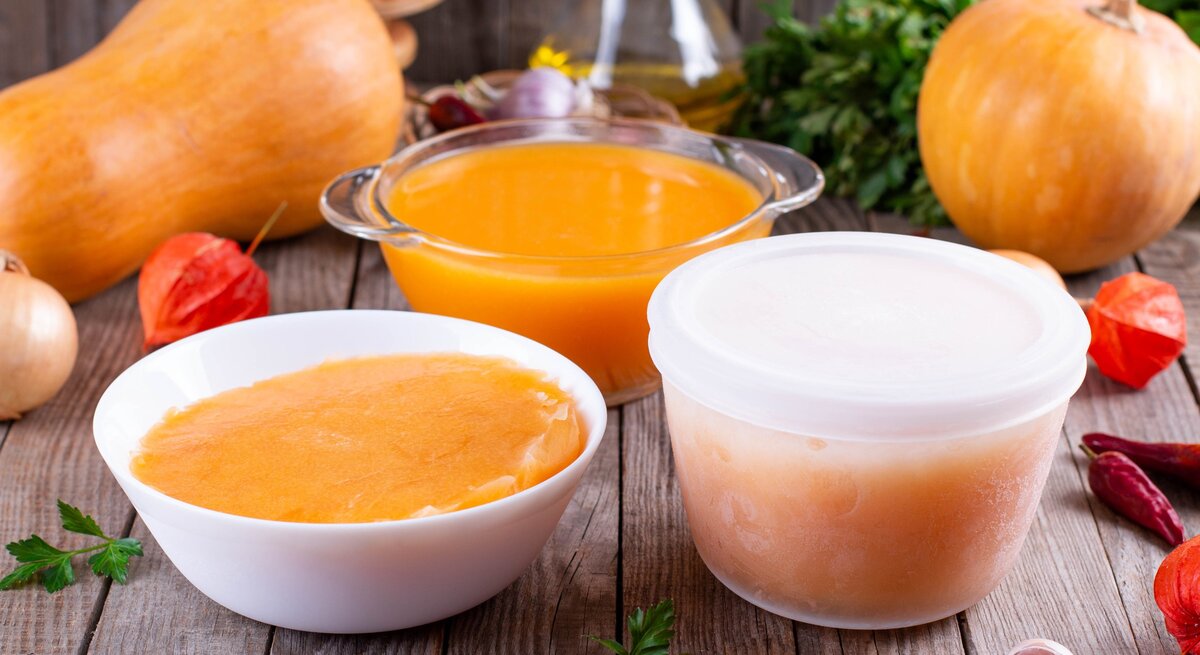 Kürbissuppe einfrieren Kürbissuppe in Gefäßen auf Holztisch. Im Hintergrund eine Karaffe mit Öl, Kürbisse und Zutaten. Frontalansicht.