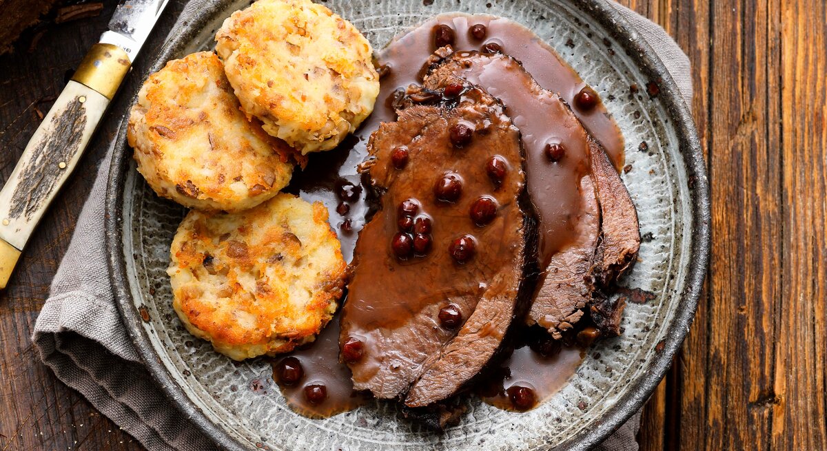 Ein Teller auf dem der Wilde Sauerbraten in Lebkuchensoße mit Kartoffeln-Maronen-Münzen angerichtet ist, neben einem Messer, dem restlichen, angeschnittenen Braten, auf einem Tuch vor einem Holzhintergrund, von oben. 