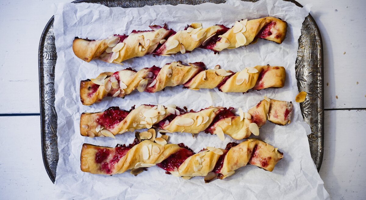 Auf einem Tablett liegen vier Rollen Blätterteigstangen mit Marmelade. Sie sind mit Mandelblättchen verziert.