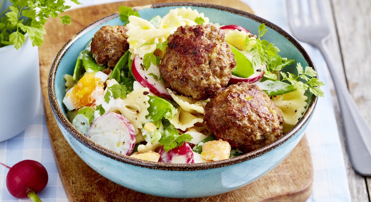 Buletten mit Nudelsalat, bestehend aus gekochten Eiern, Radieschen und Gurken, garniert mit frischem Kerbel und serviert in einer blauben Tonschale auf einem Holzbrett. 