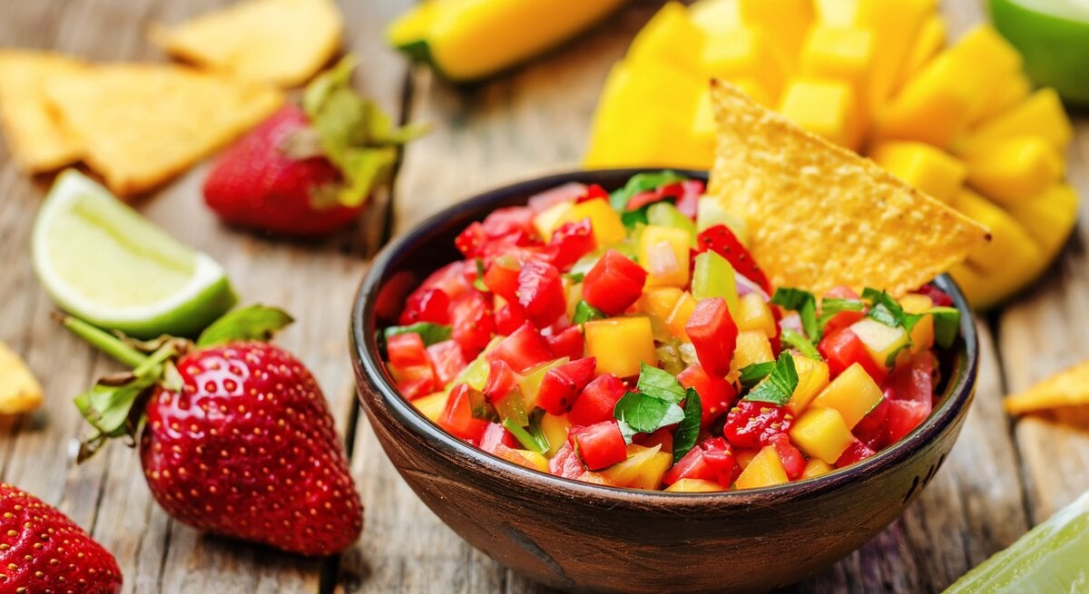 Eine Schale Erdbeer-Mango-Salsa mit Koriander, darin steckt ein Nacho-Chip. Drumherum Zutaten wie frische Mango, Erdbeeren und Limettenspalten. | © Adobe Stock/ nata_vkusidey