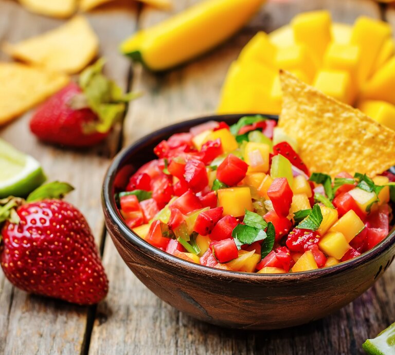 Eine Schale Erdbeer-Mango-Salsa mit Koriander, darin steckt ein Nacho-Chip. Drumherum Zutaten wie frische Mango, Erdbeeren und Limettenspalten. | © Adobe Stock/ nata_vkusidey