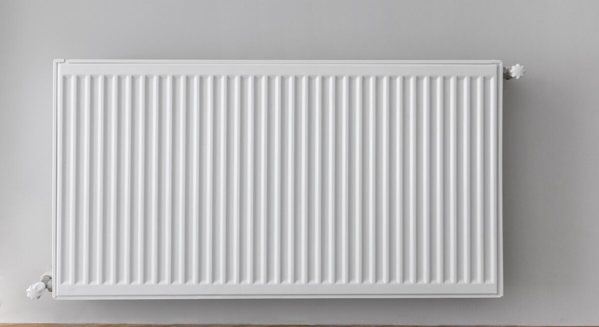 Eine weiße Heizung vor einer weißen Wand, darunter brauner Holzfußboden.