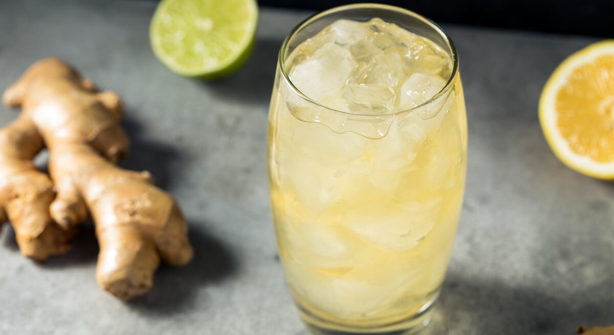 Ein Glas mit Ingwerlimonade auf einem grauen Untergrund. Daneben eine halbe Limette und eine halbe Zitrone sowie eine Ingwerknolle.