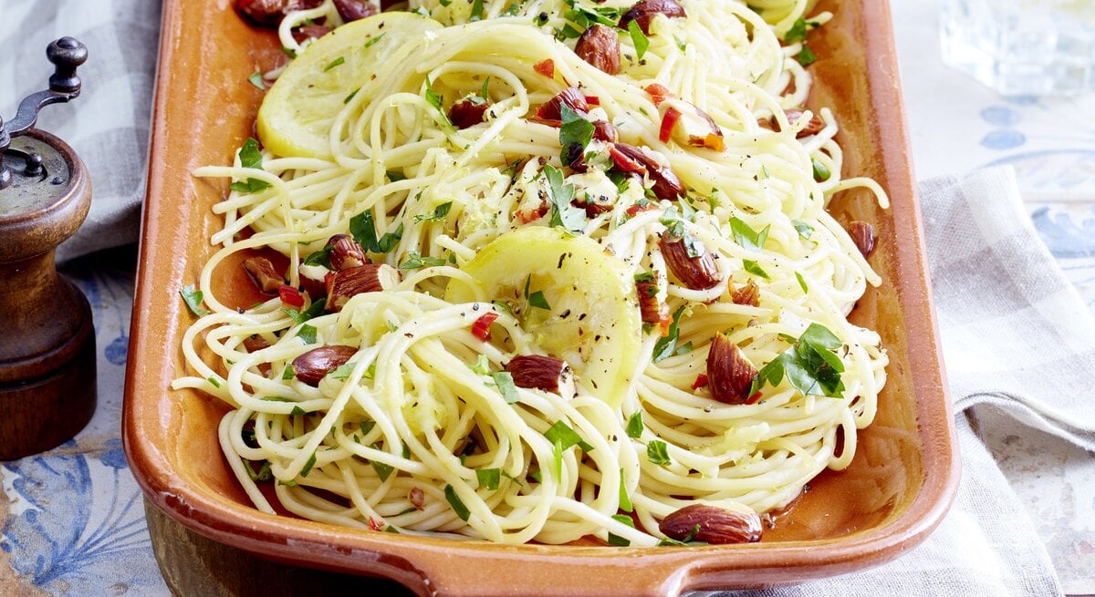 Zitronen-Mandel-Spaghetti, bestreut mit Chili und frischem Pfeffer sowie gehackter Petersilie, in einer Tonform auf gedecktem Tisch mit Tischdecke serviert. 
