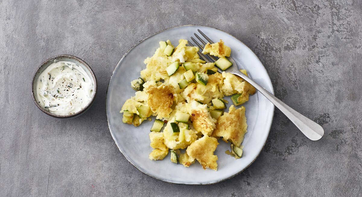 Ein heller Teller mit Halloumi-Zucchini-Schmarrn und einer Gabel steht auf einem grauen Untergrund. Links neben dem Teller steht eine kleine Schmale mit Schmarrn-Dip.