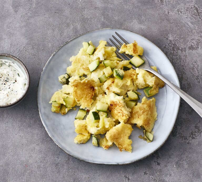 Ein heller Teller mit Halloumi-Zucchini-Schmarrn und einer Gabel steht auf einem grauen Untergrund. Links neben dem Teller steht eine kleine Schmale mit Schmarrn-Dip. | © Stockfood/ Gräfe & Unzer Verlag / Hoersch, Julia