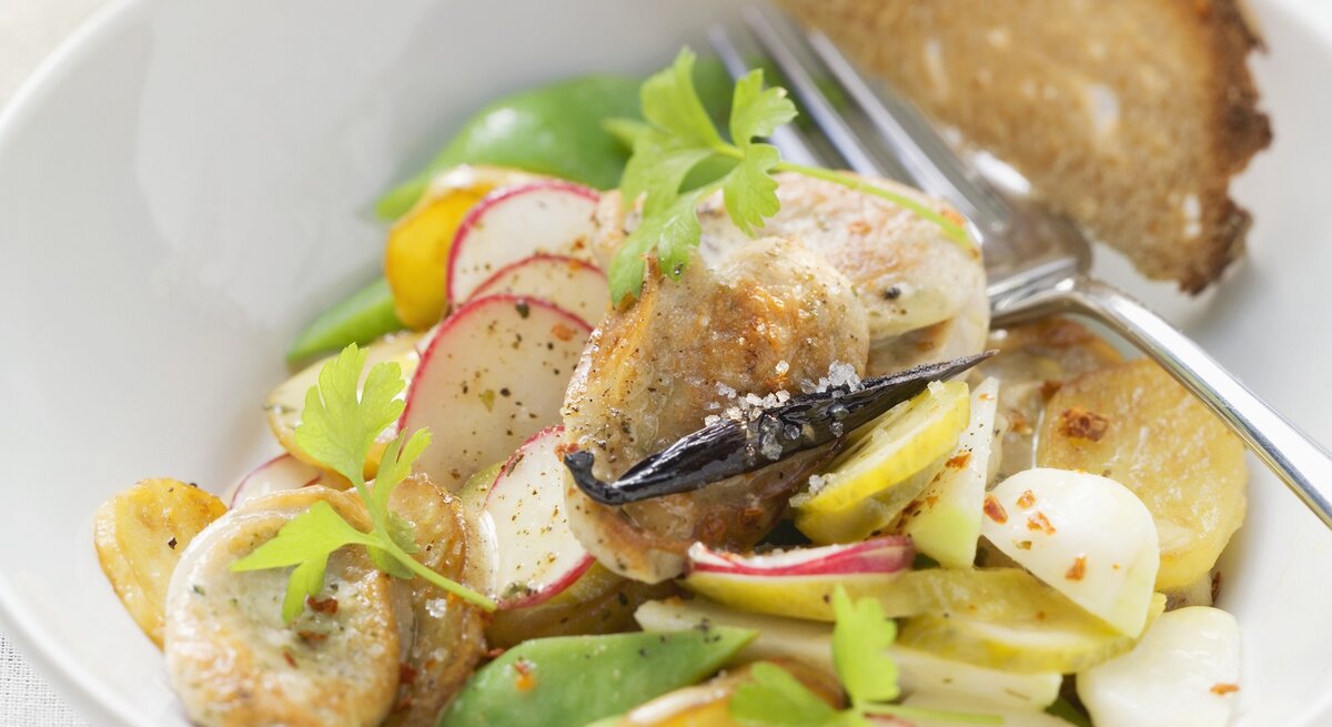 Ein weißer Tiefer Teller mit dem Bratwustsalat auf einem Tisch mit weißer Tischdecke sehr nah von schräg oben fotografiert.