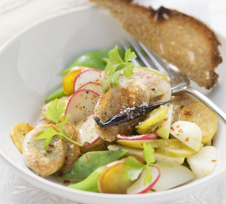 Ein weißer Tiefer Teller mit dem Bratwustsalat auf einem Tisch mit weißer Tischdecke sehr nah von schräg oben fotografiert. | © StockFood / Eising Studio - Food Photo & Video