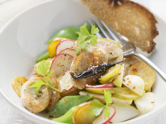 Ein weißer Tiefer Teller mit dem Bratwustsalat auf einem Tisch mit weißer Tischdecke sehr nah von schräg oben fotografiert. | © StockFood / Eising Studio - Food Photo & Video