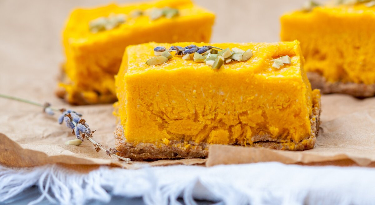 Drei Stücke von dem Kokos-Kürbis-Cheesecake auf Backpapier und einem grauen Tisch. Sehr nah von der Seite fotografiert.