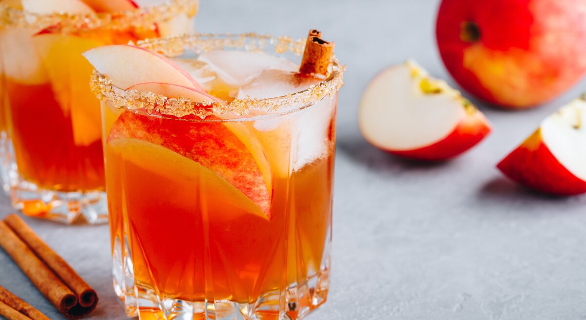 Zwei Gläser weihnachtliche Margarita mit Apfelspalten, Zimtstangen und Eiswürfeln. Daneben liegt ein angeschnittener Apfel.