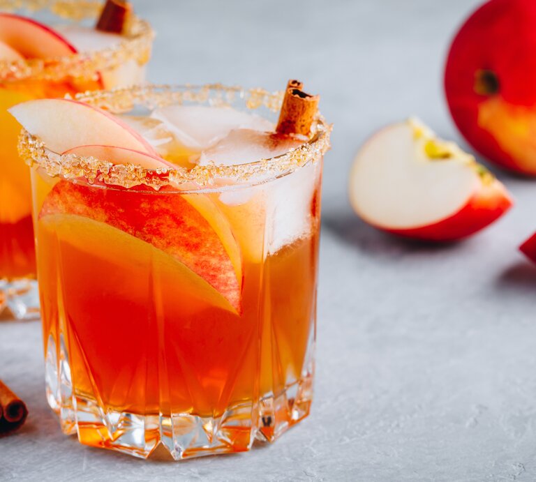 Zwei Gläser weihnachtliche Margarita mit Apfelspalten, Zimtstangen und Eiswürfeln. Daneben liegt ein angeschnittener Apfel. | © Getty Images/ wmaster890