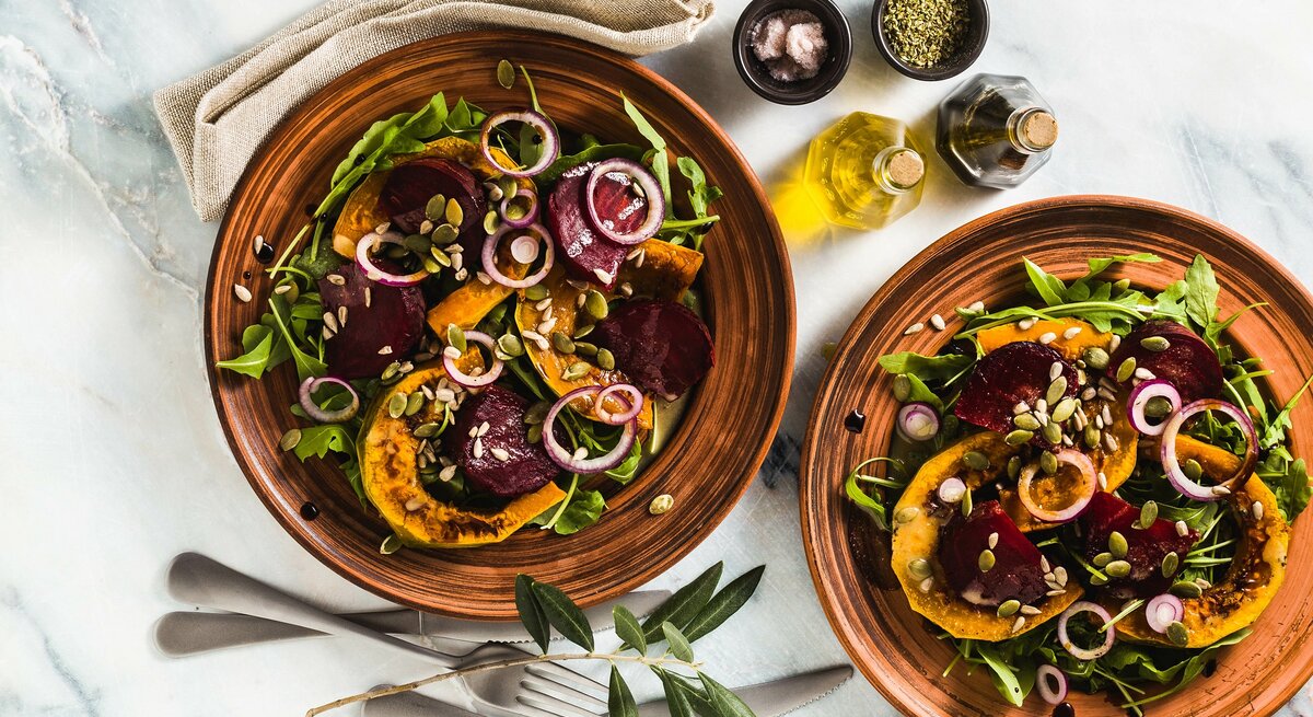 Zwei Teller Ofensalat mit Kürbis und Rote Bete sowie grünem Salat und Zwiebelringen, Draufsicht, daneben zwei Schalen mit Gewürzen und je eine Essig- und Ölflasche sowie eine Stoffserviette und Besteck.