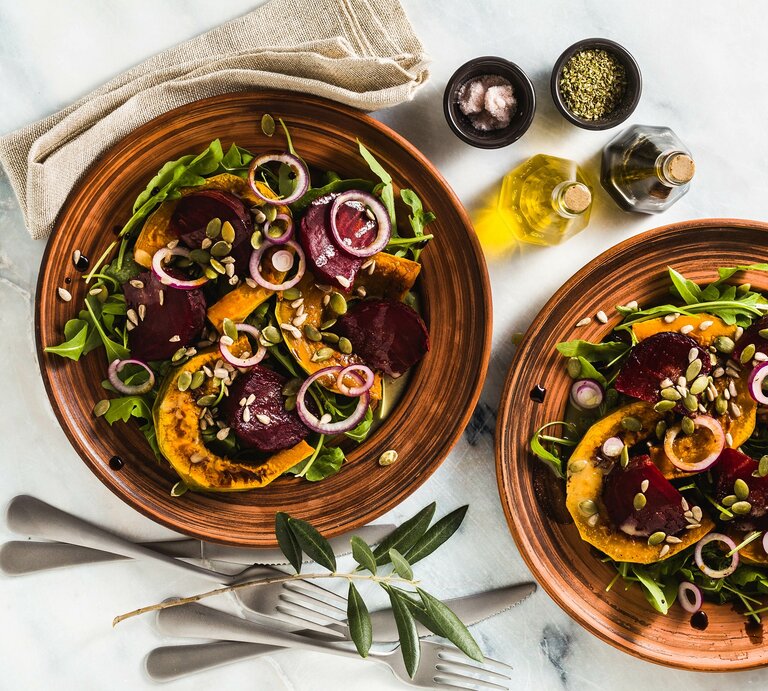 Zwei Teller Ofensalat mit Kürbis und Rote Bete sowie grünem Salat und Zwiebelringen, Draufsicht, daneben zwei Schalen mit Gewürzen und je eine Essig- und Ölflasche sowie eine Stoffserviette und Besteck. | © Adobe Stock/ irinagrigorii