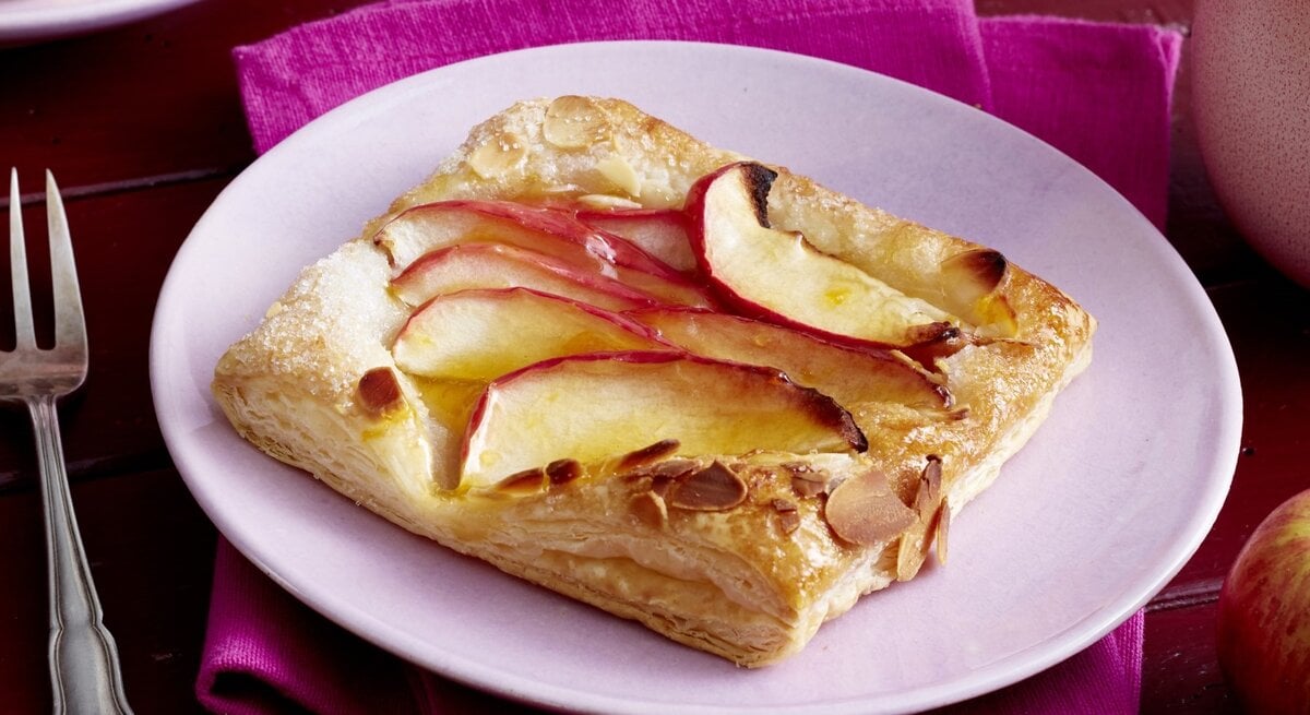 Auf einem lila Teller liegt ein Apfel-Knusper-Teilchen. Es ist goldbraun gebacken und sieht knusprig aus.