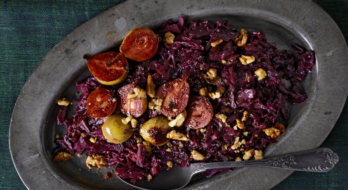 Eine ovale Platte auf der das geschmorte Rotkohl mit Birnen mit Walnüssen garniert und einem Löffel serviert wird, vor einem dunkelgrünen Hintergrund, von oben. 