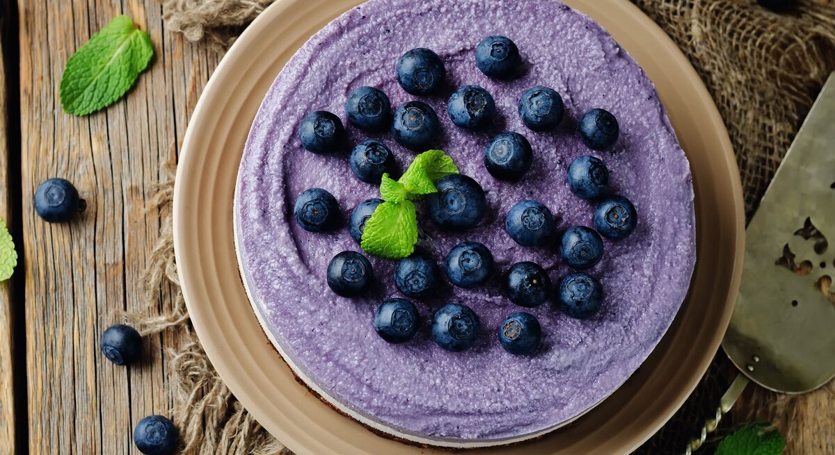 Auf einem Holztisch steht ein Heidelbeer-Käsekuchen ohne Backen. Er ist mit Blaubeeren dekoriert. die Creme ist lila eingefärbt.