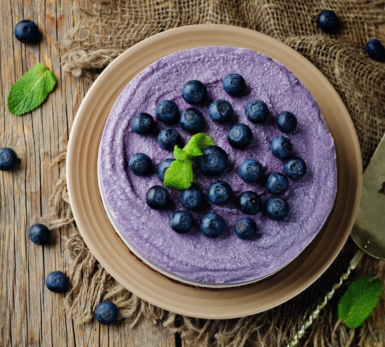 Auf einem Holztisch steht ein Heidelbeer-Käsekuchen ohne Backen. Er ist mit Blaubeeren dekoriert. die Creme ist lila eingefärbt. | © Shutterstock / Nataliya Arzamasova
