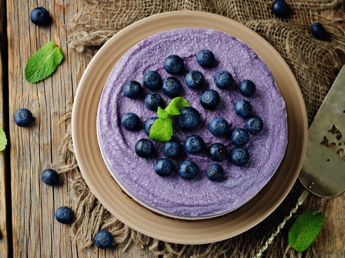 Auf einem Holztisch steht ein Heidelbeer-Käsekuchen ohne Backen. Er ist mit Blaubeeren dekoriert. die Creme ist lila eingefärbt. | © Shutterstock / Nataliya Arzamasova