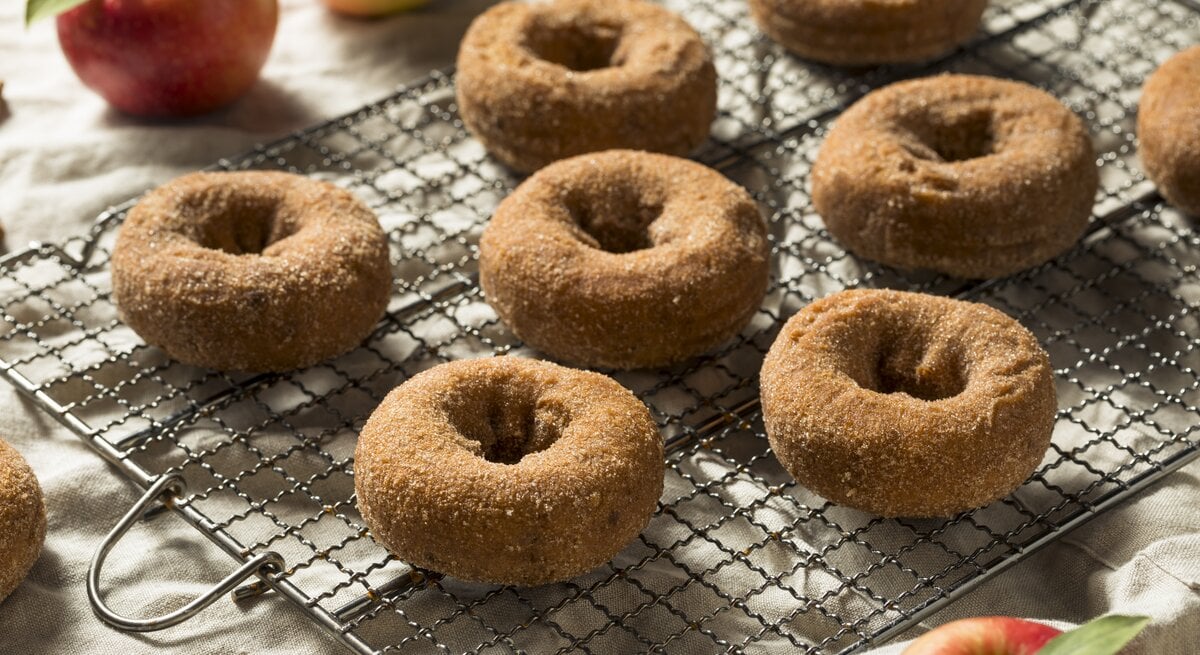 Apfelwein Donuts mit Zimt auf einem Gitter neben frischen Äpfeln auf einer hellen Tischdecke. 