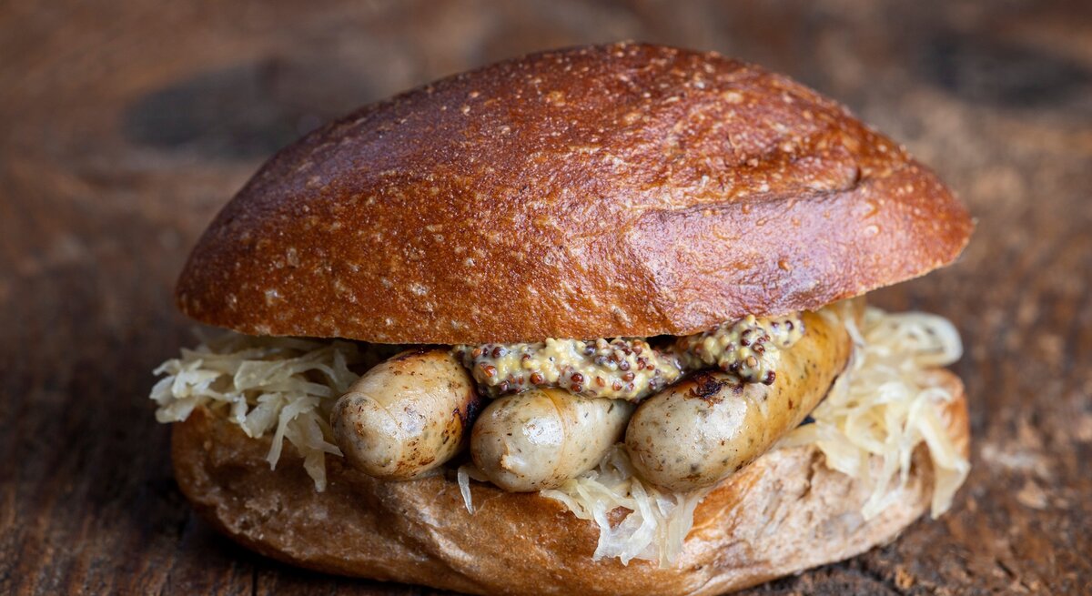 Frontal: Der fränkischer Wurstklassiker "Drei im Weggla" auf einem rustikalen Holzboden. Es handelt sich um drei Bratwürste im Brötchen mit Sauerkraut und Senf.